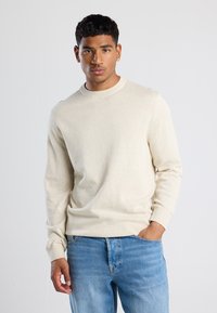 Maglione beige a maniche lunghe con scollatura rotonda e polsini a costine, realizzato in un materiale morbido, abbinato a jeans azzurri chiari.