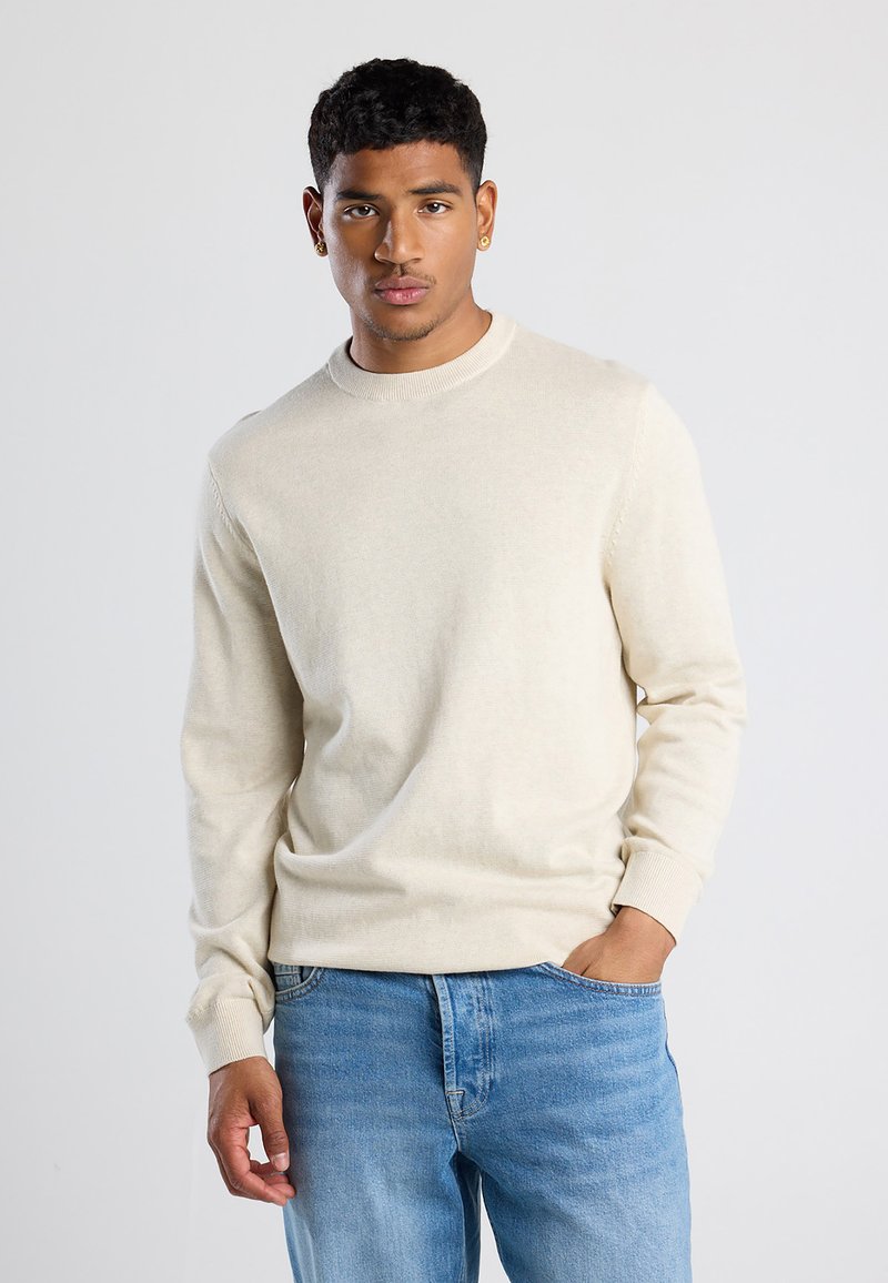 Maglione beige a maniche lunghe con scollatura rotonda e polsini a costine, realizzato in un materiale morbido, abbinato a jeans azzurri chiari.