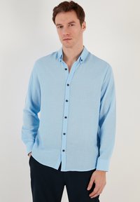 Camicia azzurro chiaro a maniche lunghe realizzata in tessuto strutturato, con chiusura a bottoni sul davanti e bottoni circolari scuri, dotata di colletto a punta.