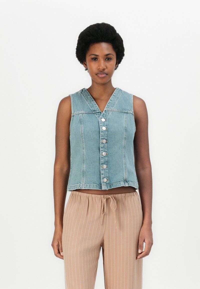 Lys blå, kort denimvest med V-hals, knappelukking og synlige sømdetaljer. Kombinert med beige stripete, løstsittende bukser.