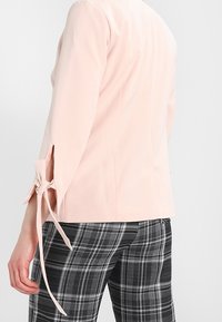 Blouse rose clair à manches trois-quarts avec liens latéraux, associée à un pantalon à carreaux noir et blanc. Tissu lisse et coupe ajustée.