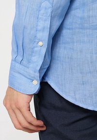Camicia da uomo in lino azzurro chiaro con polsini a bottone, caratterizzata da una texture liscia e dettagli di cucitura sottili, abbinata a pantaloni scuri e con texture.