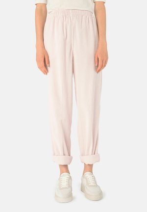 Vrouw draagt lichtroze gestreepte losse broek met opgerolde zoom en witte sneakers, staand tegen een effen achtergrond.