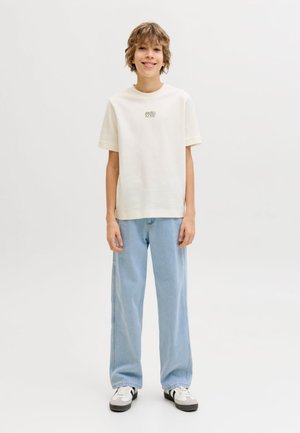 Joven sonriente con camiseta blanca de manga corta con texto, vaqueros azul claro y zapatillas blancas con detalles negros, de pie.