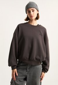 Brown oversized sweatshirt with dropped shoulders and ribbed hem, paired with gray knitted beanie. Casual look with soft fabric texture.