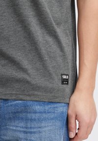 Grå T-shirt lavet af blødt materiale, med rund halsudskæring, korte ærmer og en rektangulær sort etiket med hvid tekst i sømmen.
