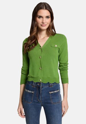 Cardigan en tricot vert avec un col en V, un ourlet festonné et des boutons dorés. Présente une poche poitrine et des manches côtelées. Porté avec un jean bleu.