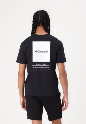 Person mit geflochtenem Haar, die ein schwarzes Columbia-T-Shirt und schwarze Shorts trägt und mit dem Rücken zur Kamera vor einem schlichten weißen Hintergrund steht.