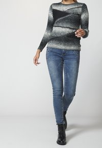 Harmaa ja musta neulottu villapaita, jossa on teksturoitu geometristen kuvioiden kuvio, yhdistettynä sinisiin farkkuihin, joissa on kukkaompeleita, ja mustiin nauhallisiin saappaisiin.