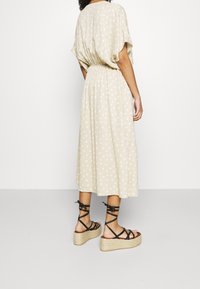 Frau in beiger Midi-Kleid mit Pünktchen und tailliertem Schnitt, trägt schwarze Schnür-Plateausandalen und steht vor weißem Hintergrund.