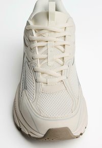 Bežové športové tenisky s prepracovanou sieťovinou, polstrovaným jazykom a tonálnymi šnúrkami. Gumená podrážka s jemnými textúrovanými detailmi.