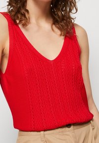Červený pletený top bez rukávů se V-výstřihem, texturovaným kabelovým vzorem a otevřeným švěm. Materiál je lehký s volným střihem.