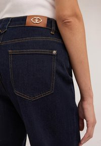 Nära bild av en person som bär mörkblå denimjeans med en brun läderpatch och metall-logotyp på midjebandet, som visar deras högra arm.
