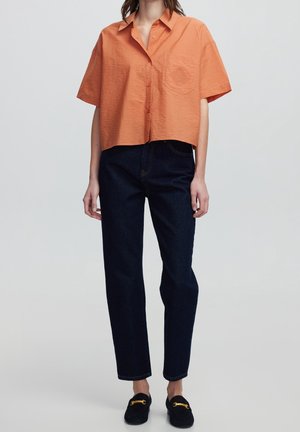 Pomarańczowa skrócona koszula z krótkim rękawem z detalem kieszeni, zestawiona z ciemnymi, prostymi jeansami i czarnymi mokasynami z złotymi akcentami.