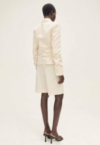 Blazer en lin beige avec des épaules structurées, associé à un short assorti. Des sandales à talons noires complètent le look. Vue arrière, cadre minimal.