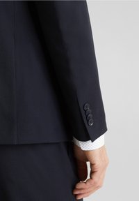 Manica di giacca navy con quattro bottoni scuri, vestibilità su misura, con il polsino di una camicia a pois chiari visibile sotto. Texture liscia.