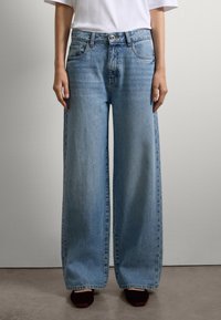 Persona che indossa jeans larghi blu chiaro, una maglietta bianca a maniche corte e scarpe nere a punta, in piedi contro un muro semplice.