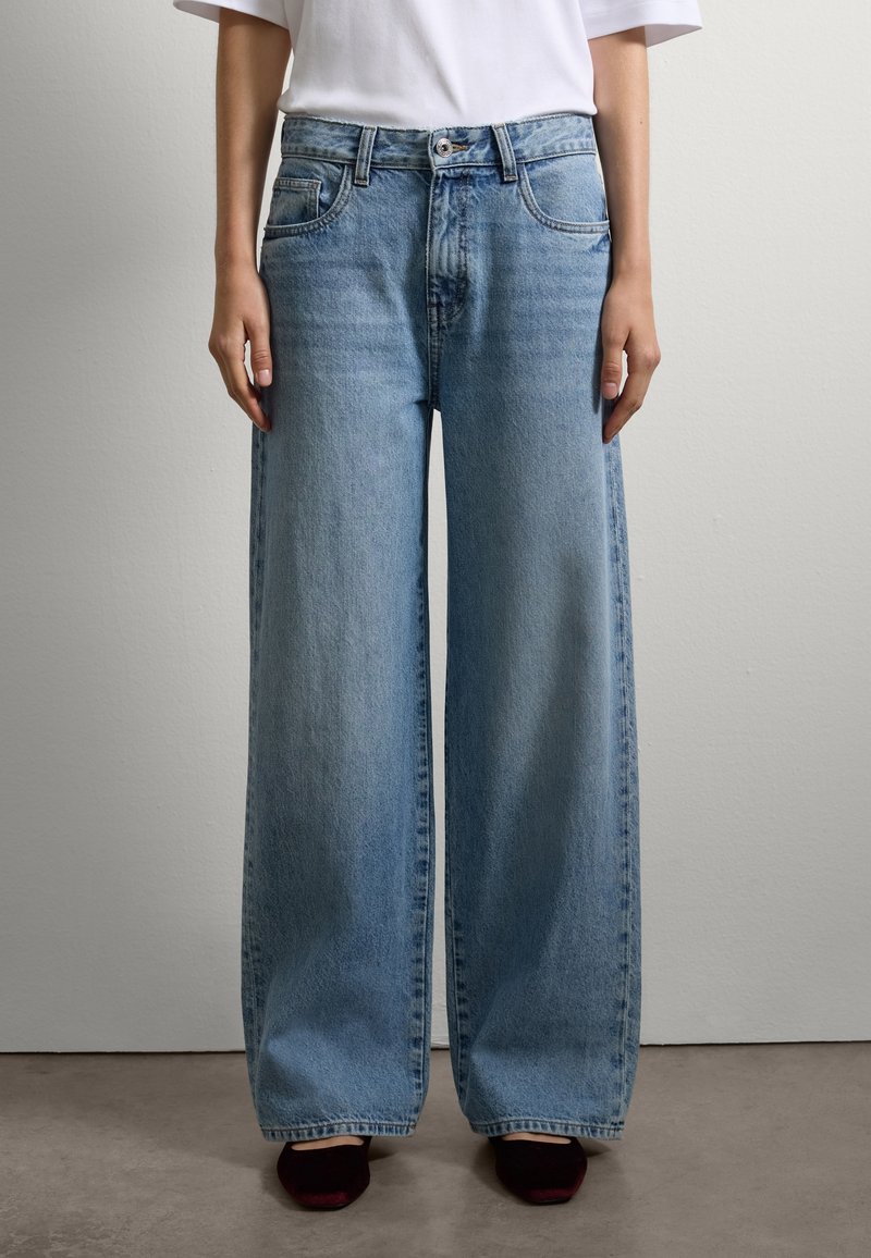 Persona che indossa jeans larghi blu chiaro, una maglietta bianca a maniche corte e scarpe nere a punta, in piedi contro un muro semplice.