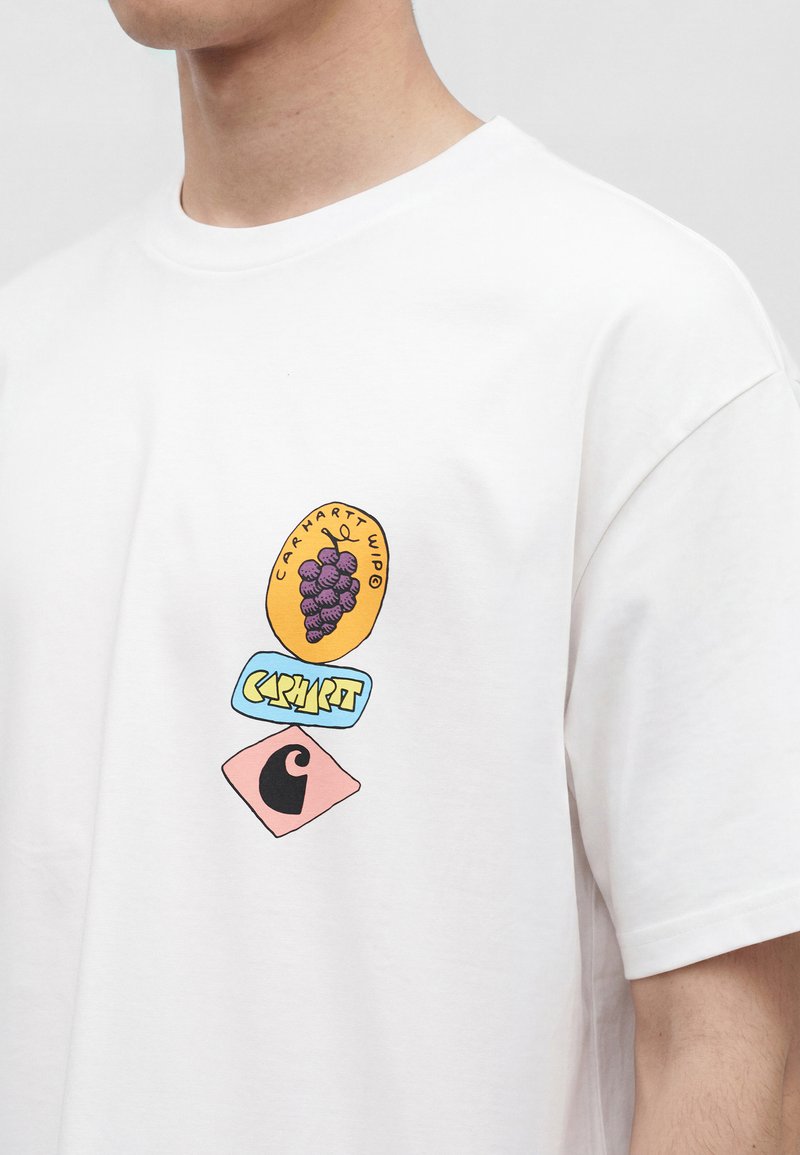 Person trägt weißes T-Shirt mit farbiger Grafik von Trauben und geometrischen Formen, einschließlich des Texts "Carhartt WIP" auf der Brust.