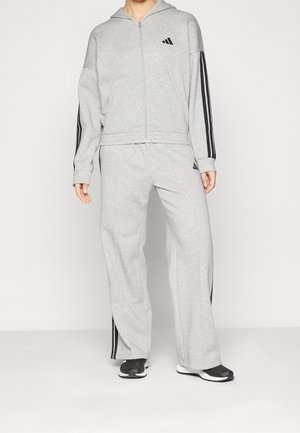 Persoon draagt een grijs Adidas trainingspak met zwarte strepen op de mouwen en broek, gecombineerd met zwart-witte sneakers.