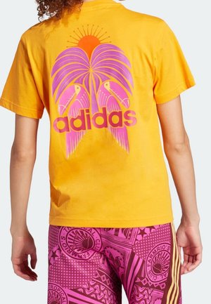 Personne portant un t-shirt jaune Adidas avec un motif d'oiseau tropical rose et un soleil, assorti à un short rose à motifs avec divers logos.