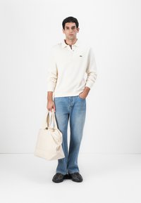 Krämfärgad tröja, lång ärm, litet grönt logotyp. Ljusblå jeans, tote bag i beige och svarta texturerade skor. Enkelt, avslappnat outfit.