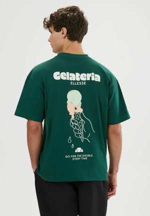 Jeune homme portant un T-shirt vert foncé avec un motif d’une glace à deux boules fondante et le texte « Gelateria » et « Prenez le double, à chaque fois. »