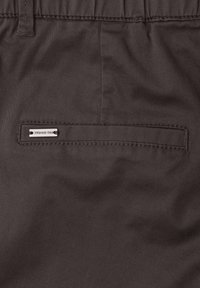 Poche arrière d’un pantalon marron avec une fente horizontale et une petite étiquette métallique rectangulaire de marque indiquant « Street One » en haut à gauche.