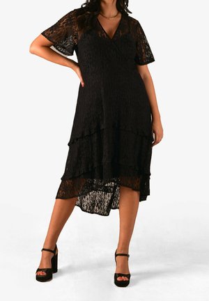 Frau trägt ein schwarzes, mehrlagiges Spitzen-Midikleid mit kurzen Ärmeln und schwarze Sandalen mit hohen Absätzen und steht mit der Hand in der Hüfte.