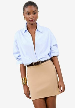 Cămașă oversized de culoare albastru deschis cu nasturi, fustă mini bej ajustată, curea neagră cu cataramă, brățară aurie la încheietura mâinii. Texturi de țesătură netedă.