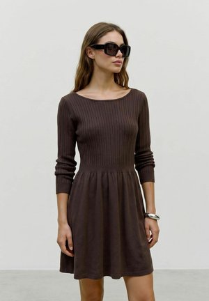 Donna con lunghi capelli castani che indossa grandi occhiali da sole neri, un vestito marrone a coste a maniche lunghe e un braccialetto d'argento, in piedi davanti a uno sfondo semplice.