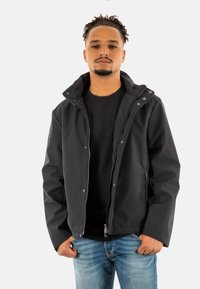 Giacca nera impermeabile con chiusura a zip, bottoni a pressione visibili e cappuccio. Indossata sopra una maglietta nera, abbinata a jeans azzurri chiari.