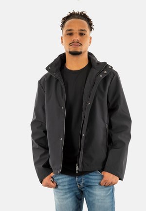 Giacca nera impermeabile con chiusura a zip, bottoni a pressione visibili e cappuccio. Indossata sopra una maglietta nera, abbinata a jeans azzurri chiari.