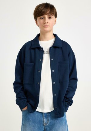 Ragazzo adolescente con capelli castani corti che indossa una giacca blu navy con bottoni sopra una t-shirt bianca stampata e jeans blu, mani in tasca.