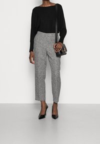 Suéter negro, pantalones grises con estampado y zapatos de tacón negro. Modelo sosteniendo un pequeño bolso negro, de pie sobre un suelo gris.