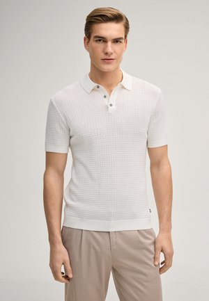 Young man standing, wearing a textured white short-sleeve polo shirt and beige pants, neutral background.