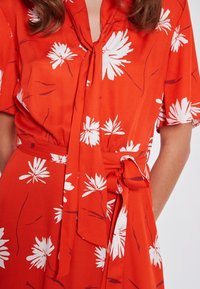 Robe fleurie rouge avec manches courtes, ceinture à nouer à la taille, et un motif de fleurs blanches avec des accents rouges subtils sur un tissu lisse.