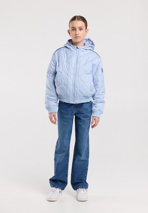 Jeune personne debout, portant une veste matelassée à capuche bleu clair, un jean bleu avec des boutons latéraux, et des baskets blanches.