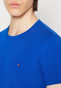 Blå bomullströja med rund halsringning. Har en liten, broderad logotyp i röd, vit och blå på bröstet. Slät textur.