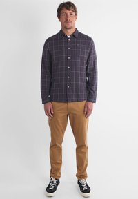 Chemise à manches longues en tartan violet foncé avec des lignes vertes, associée à un pantalon beige. Des baskets noires avec des semelles et des lacets blancs complètent la tenue.
