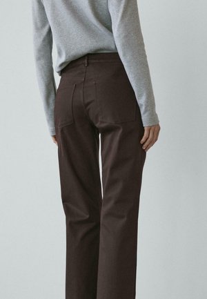 Pantaloni marrone scuro a vita alta con gamba dritta, dotati di tasche posteriori e una texture di tessuto liscia, abbinati a una camicia grigia a manica lunga.