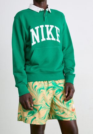 Sudadera verde de Nike con logo blanco, que presenta un cuello con solapa. Combinada con pantalones cortos amarillos que exhiben un patrón de mármol verde.