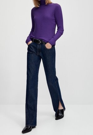 Personne portant un pull à col roulé côtelé violet, un jean bleu foncé avec des fentes latérales, une ceinture noire et des chaussures noires pointues, mains dans les poches.