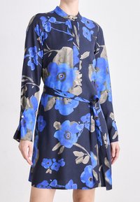 Kortärmad klänning i marinblått tyg med stora blå blommönster. Inkluderar ett bälte och knappdetaljer på ärmarna.