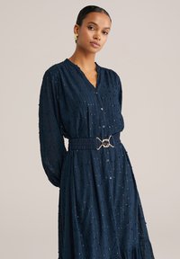 Marineblaues Hemdkleid aus strukturiertem Stoff, mit langen Ärmeln und tailliertem Schnitt, das einen kreisförmigen goldenen Gürtelverschluss aufweist. Dezente dekorative Elemente sind sichtbar.