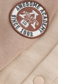 Brąska, haftowana okrągła naszywka z napisem "Awesome Academy" oraz "Athletics NYC" na beżowym płótnie, z detalem guzika i teksturowaną powierzchnią.