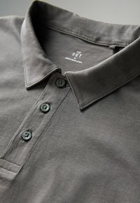 Tumši pelēks polo krekls ar pogu aizdari, kurā ir trīs pogas, mīksta tekstūra un apkakle. Etiķete norāda "THE SET, MADE IN BANGLADESH."