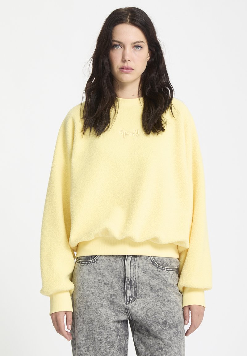 Geel, zachte fleece trui met een ronde halslijn en een geribbelde zoom. Heeft losse lange mouwen en subtiele geborduurde details.