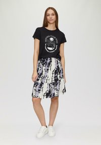 Schwarzes Baumwoll-T-Shirt mit Grafikdruck, kombiniert mit einem schichtweisen Rock in einem Batikmuster in den Farben Lila, Schwarz und Creme. Weiße Sneakers.