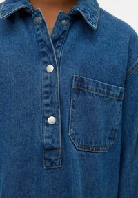 Artimas džinsinio marškinio vaizdas su sidabro spalvos snap mygtukais ir priekiniais krūtinės kišenėmis, rodantis mėlynos medžiagos tekstūrą ir oranžinius siūlus.